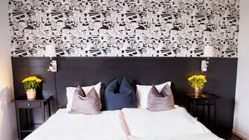A bedroom with a patterned wallpaper, a bed with pillows, two bedside tables with lamps, and a nightstand with a flower vase.