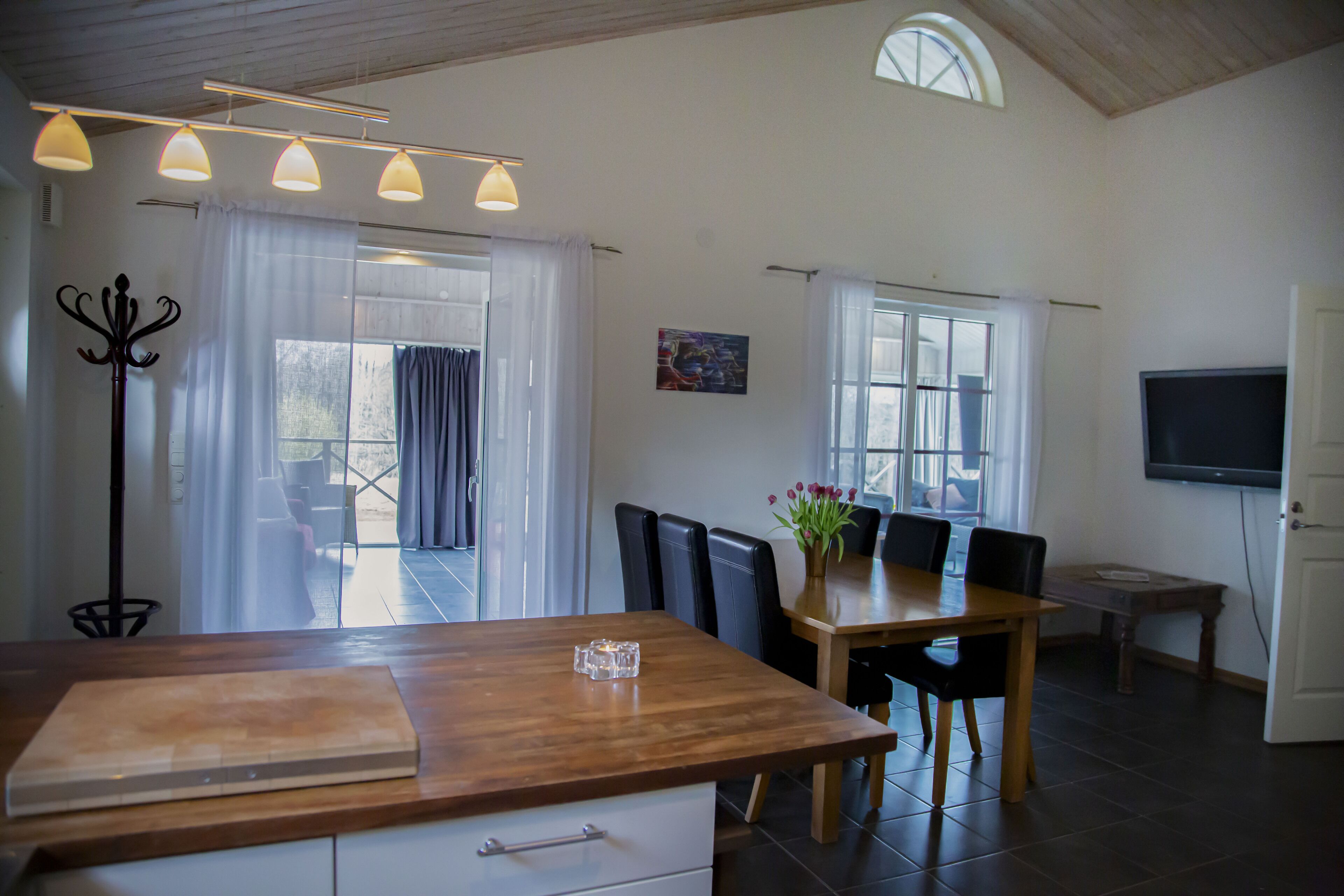 Una cocina moderna con encimera de madera, comedor con sillas negras, televisor montado en la pared y un ventanal con cortinas.