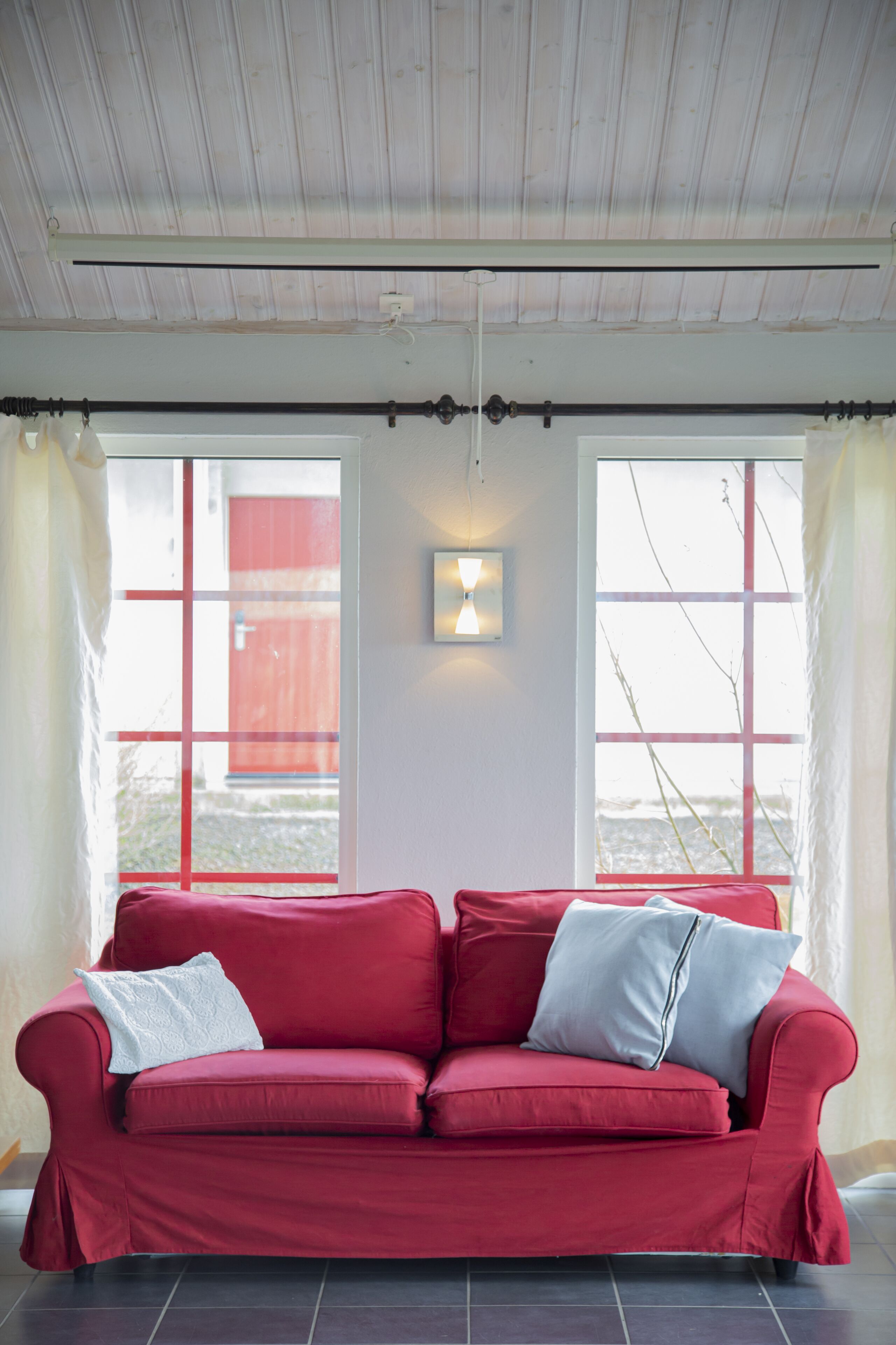 A red sofa with two pillows, a wall-mounted light fixture, and a window with a view of a red building.
