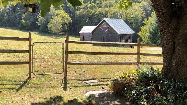 A rustic barn with a metal roof and a colorful diamond-shaped emblem, surrounded by a wooden fence and lush greenery. - Relax in a Peaceful Country Home in the Blue Ridge Mountains. Enjoy Local Events (Sparta)