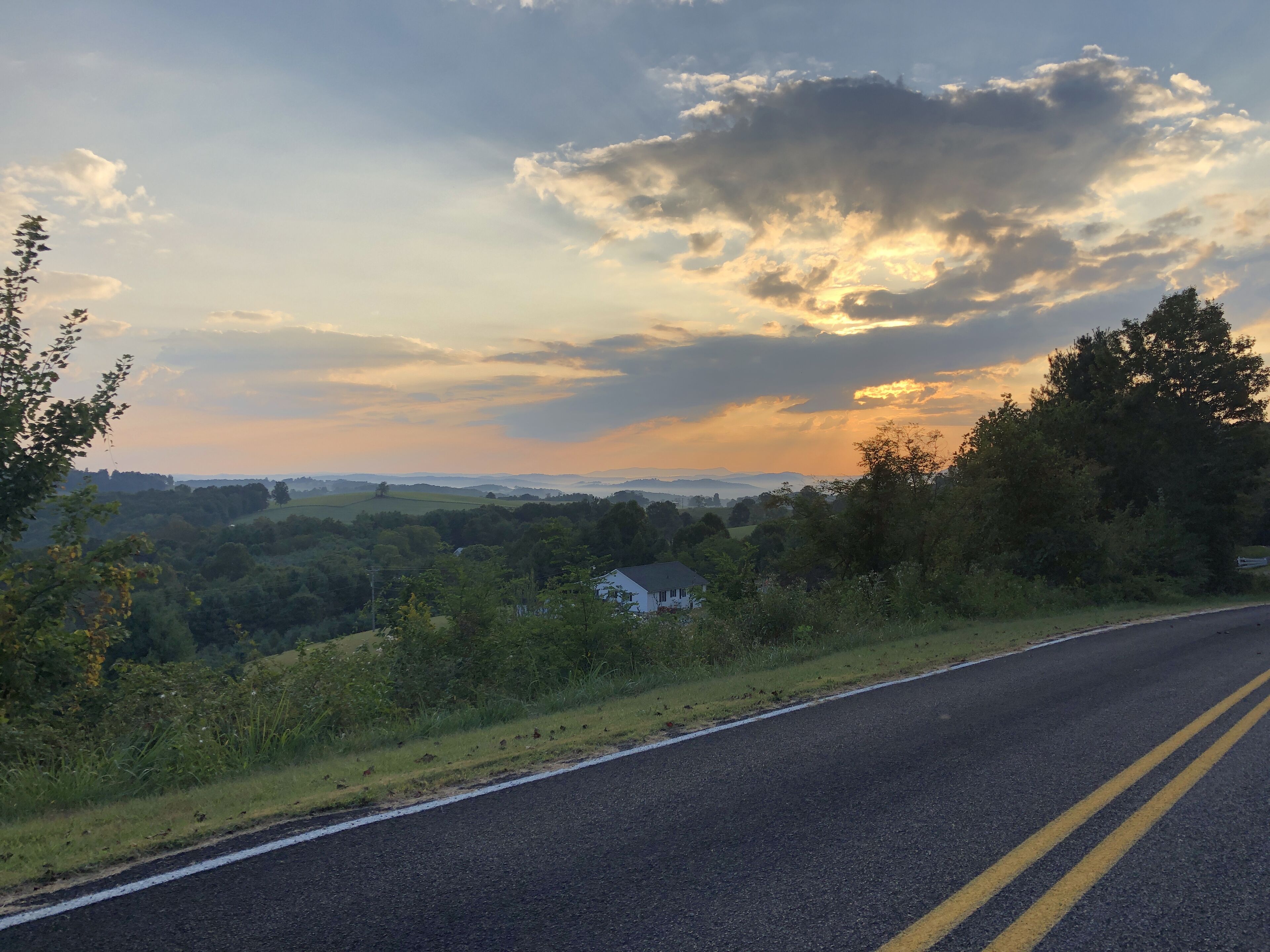 A rural road with a house on the side, surrounded by greenery and a cloudy sky.