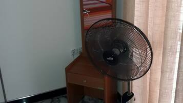 A room with a bed, a wooden nightstand, a standing fan, and a mirror.