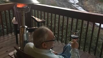 A man seated on a wooden deck chair, holding a glass of wine, with a view of a waterfall in the background.