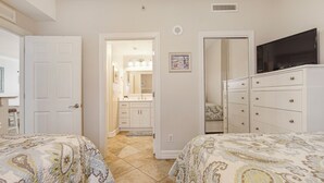 A bedroom with two beds, a dresser, a television, and a bathroom visible through an open door. - Tropic Winds 1504 - Amazing beachfront condo! Beach chairs included! (Panama City Beach)