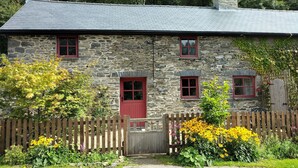Una casa de piedra con puerta y ventanas rojas, rodeada de una cerca de madera y plantas con flores.