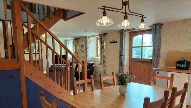 Una escalera de madera lleva a un comedor con una mesa y sillas, un florero con flores y una zona de cocina con una cesta para pan.