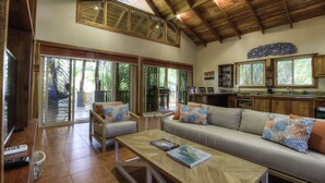 A spacious living area with a wooden ceiling, a sofa set, a coffee table, and a kitchen area with wooden cabinets and a window overlooking greenery. - Family Oasis Minutes Walk to the Playa Guiones (Nosara)