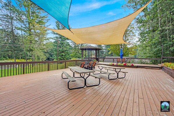 A wooden deck with picnic tables and chairs, surrounded by trees and a gazebo.