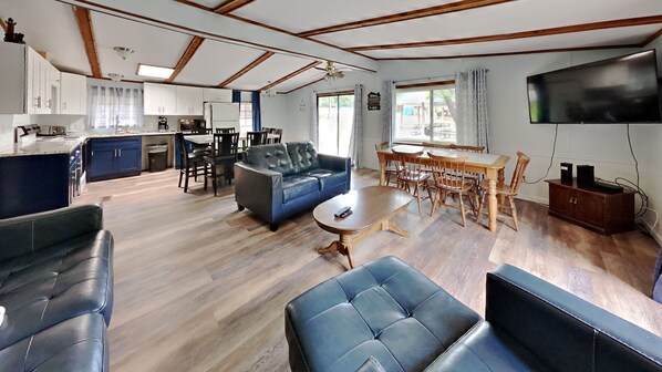 A spacious living area with a sofa, a dining table with chairs, a kitchen with a refrigerator, and a TV mounted on the wall.