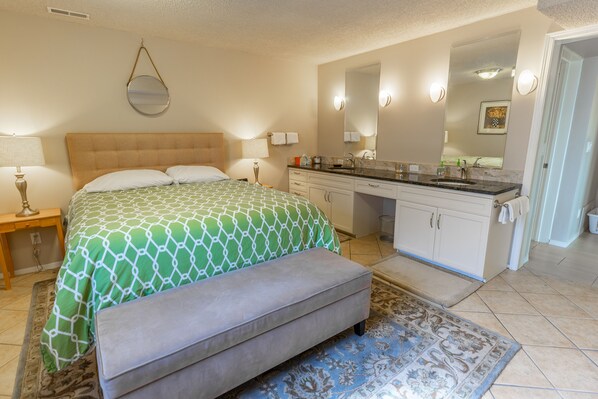 A bedroom with a bed, bedside tables, a bench, a bathroom with a sink, mirror, and cabinets.