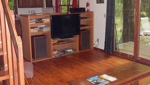 Een ruime woonkamer met een houten vloer, een televisie op een houten dressoir, een plafondventilator en een groot raam met uitzicht op het buitengebied.
