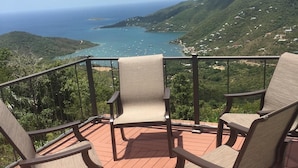 Una terraza de madera con tres sillas de mimbre orientadas hacia una vista panorámica de la costa, con barcos y vegetación.