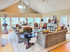 Uma sala de estar ampla com teto alto, janelas grandes e diversos móveis, incluindo sofás, uma mesa de centro e objetos decorativos.