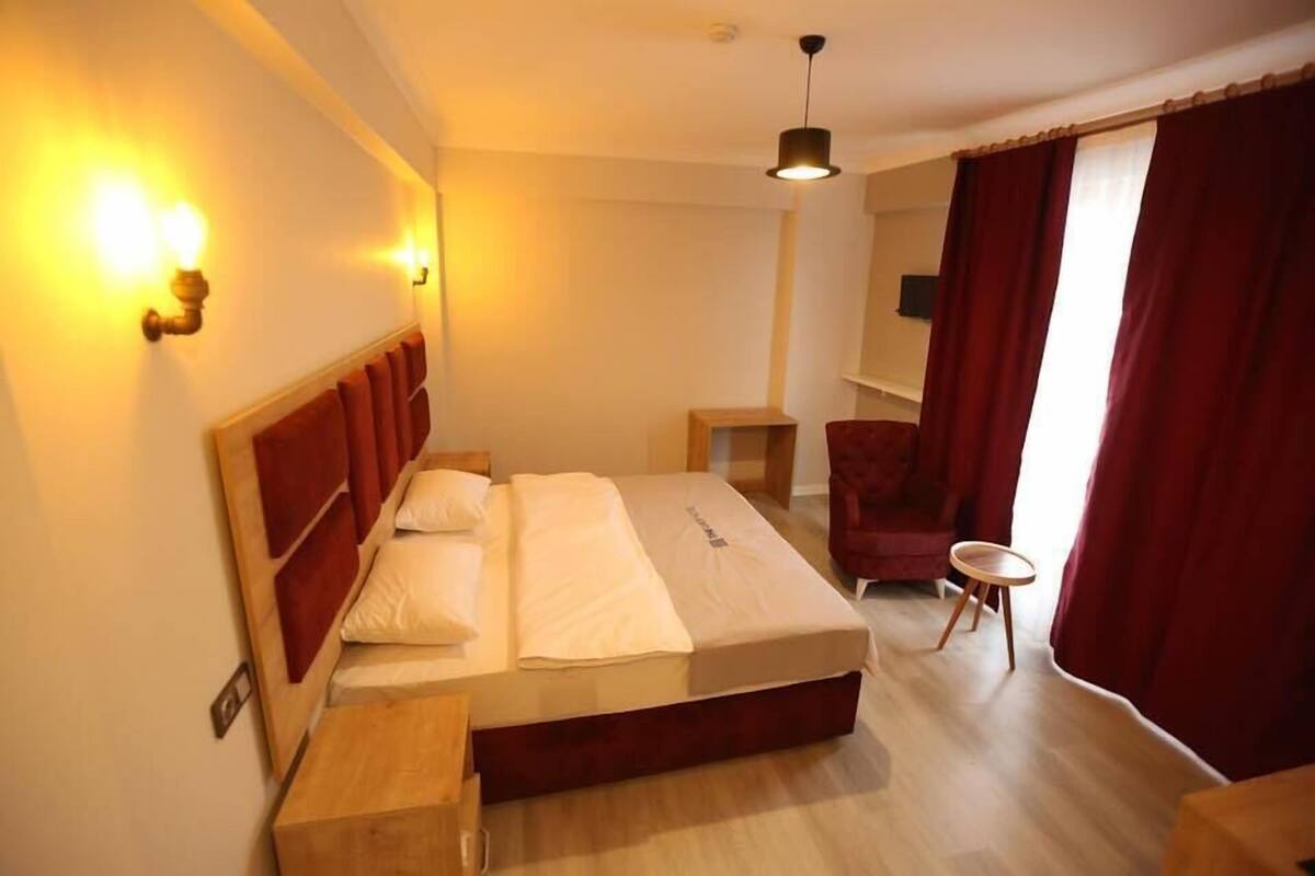 A hotel room with a large bed, a wooden nightstand, a small table, a red armchair, and a window with red curtains.