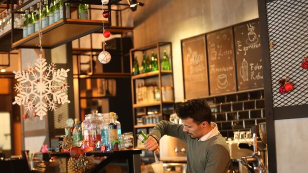 Un barista preparando una bebida en la barra, con estantes llenos de botellas y un menú en la pizarra al fondo.