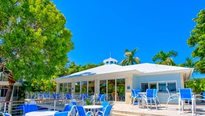 A covered outdoor seating area with blue chairs and tables, surrounded by palm trees and a clear blue sky. - Tropical Escape at Mariner’s Club  2BR Key Largo Condo w/ Pools & Marina (Key Largo)