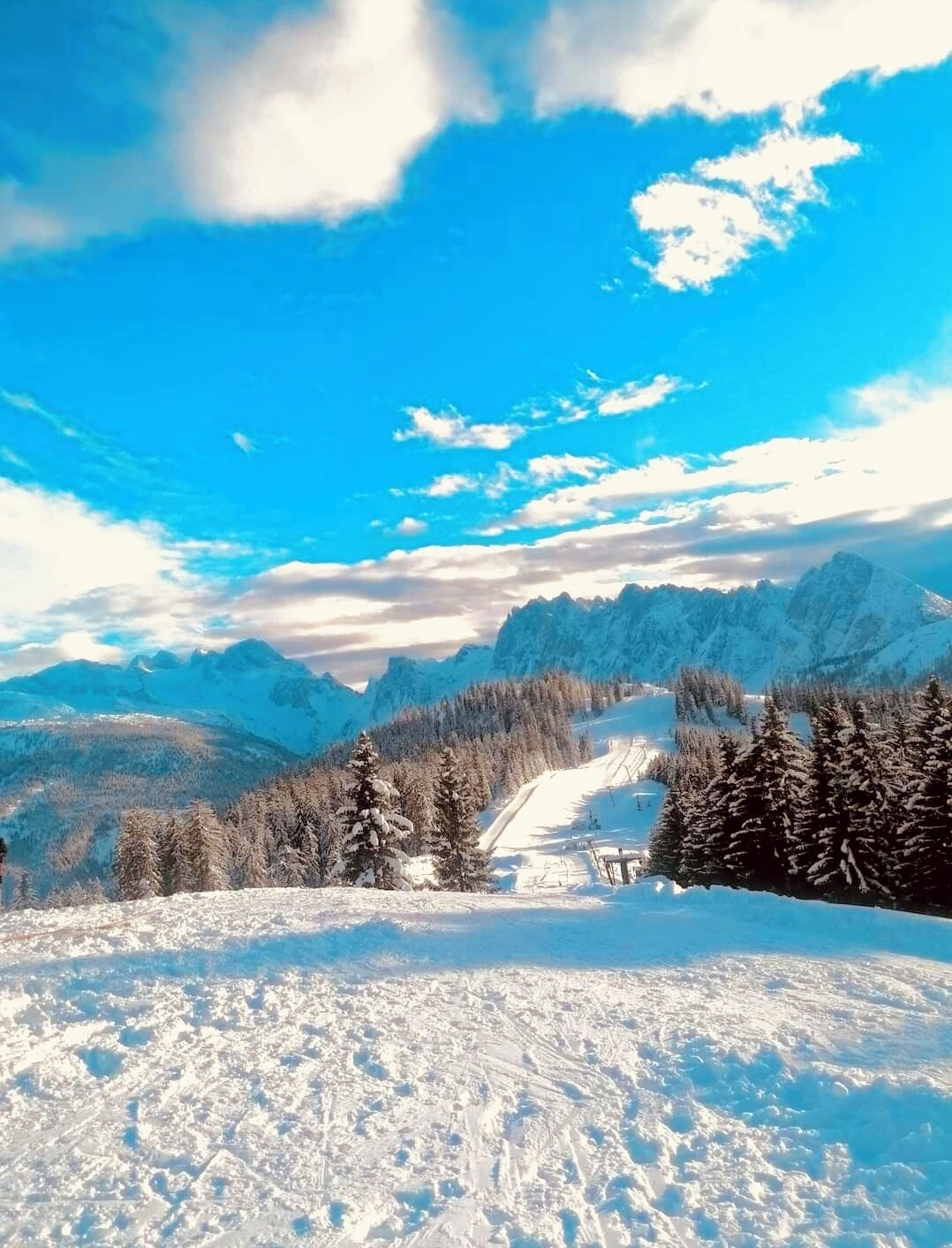 Eine verschneite Berglandschaft mit Skipiste, Bäumen und blauem Himmel.