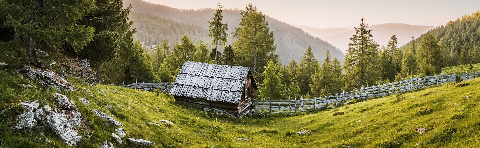 A rustic cabin with a sloped roof, surrounded by a wooden fence and dense forest.