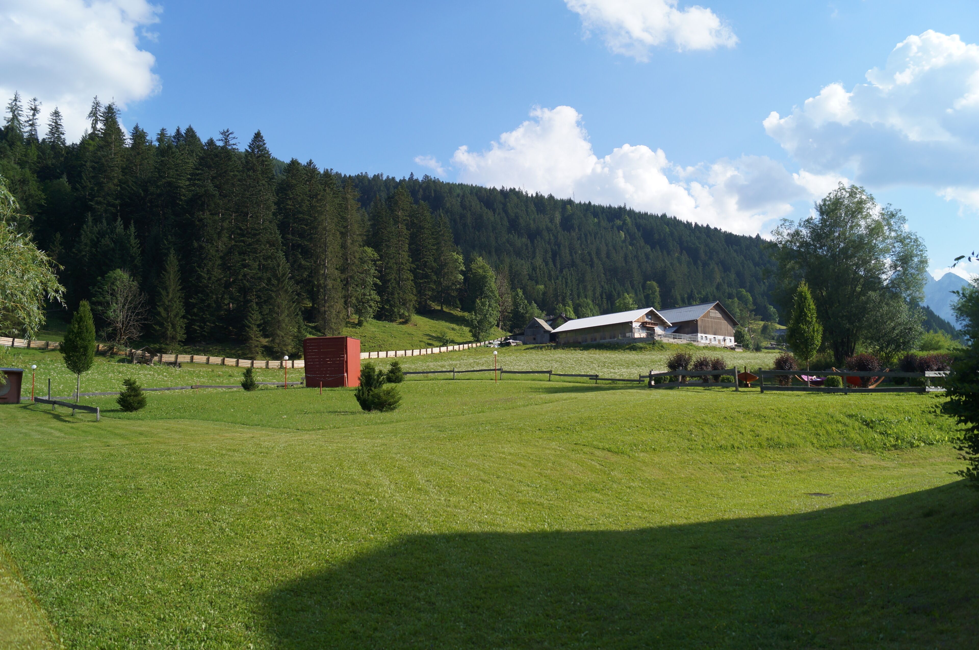Paisaje rural con una casa de campo, un campo cercado y una montaña boscosa al fondo.