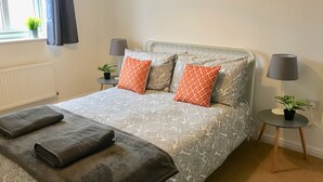 Ein ordentlich gemachtes Bett mit gemusterten Bettbezügen und zwei Kissen, ein Nachttisch mit Lampe, ein Blumentopf und Blick durch einen Vorhang drapierten Fensters.