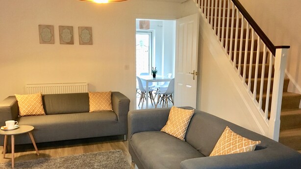 Ein Wohnzimmer mit zwei grauen Sofas, einem kleinen runden Tisch mit Tasse und Untertasse, einem grauen Teppich und einer Treppe mit einem Holnlaufgeländer.