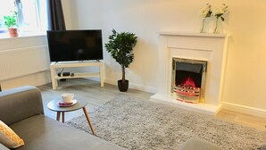 Ein Wohnzimmer mit grauem Sofa, einem Flachbildfernseher, einem Blumentopf, einem Kamin mit Feuer und einem kleinen Tisch mit einer Tasse.