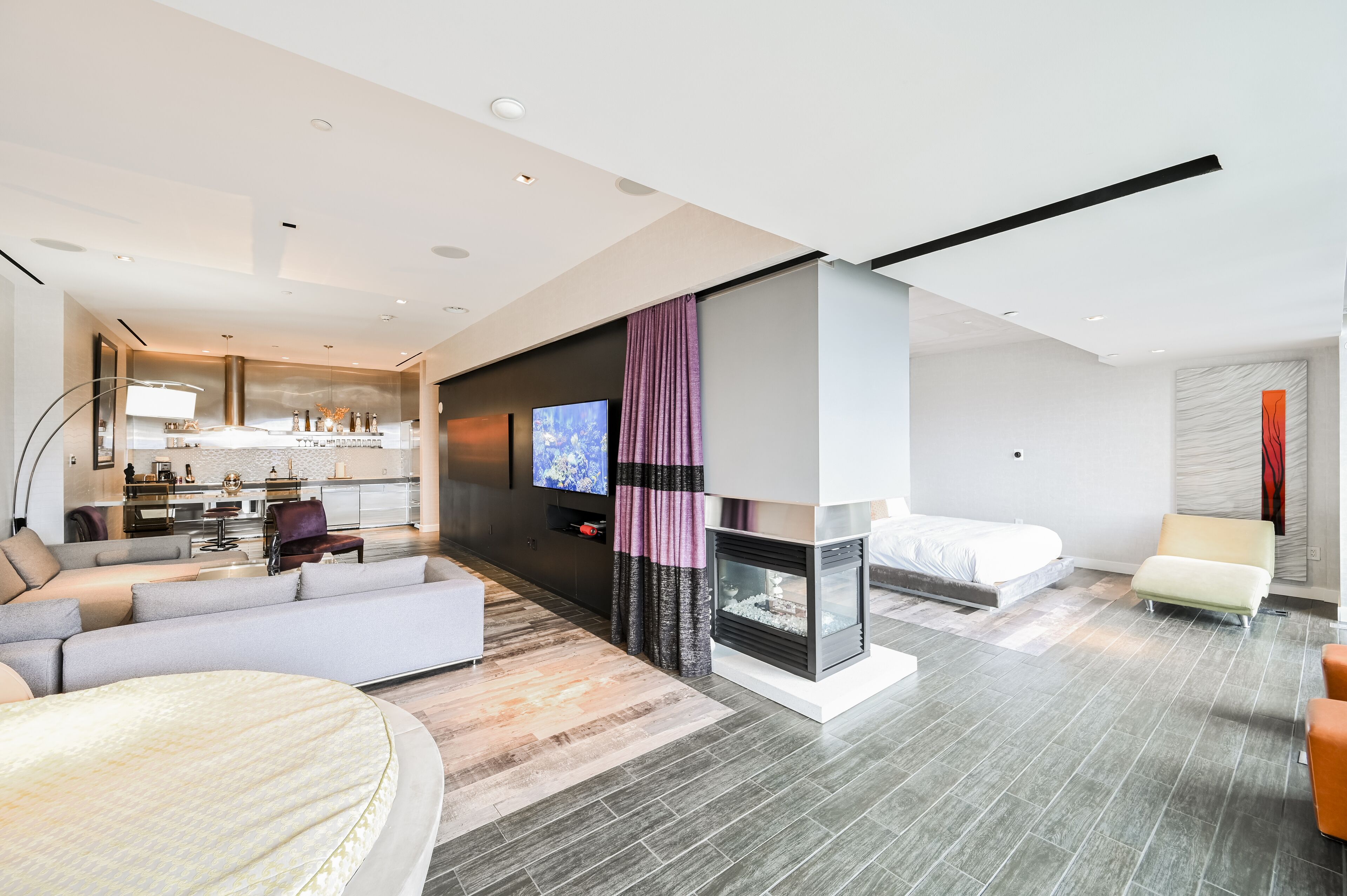 A modern hotel room with a living area, a fireplace, a bed, and a kitchen in the background.