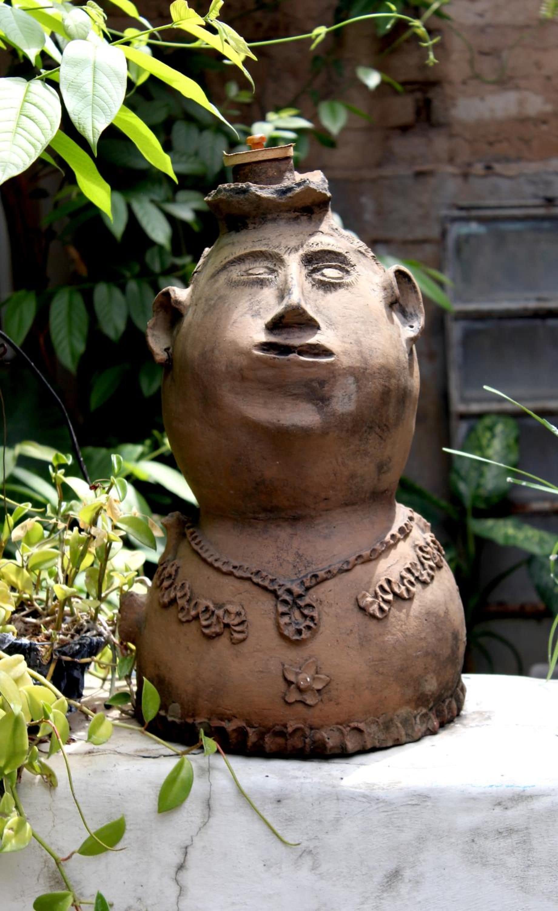 Una escultura de terracota con motivos florales y una maceta pequeña en la parte superior, sobre una superficie blanca rodeada de plantas verdes.