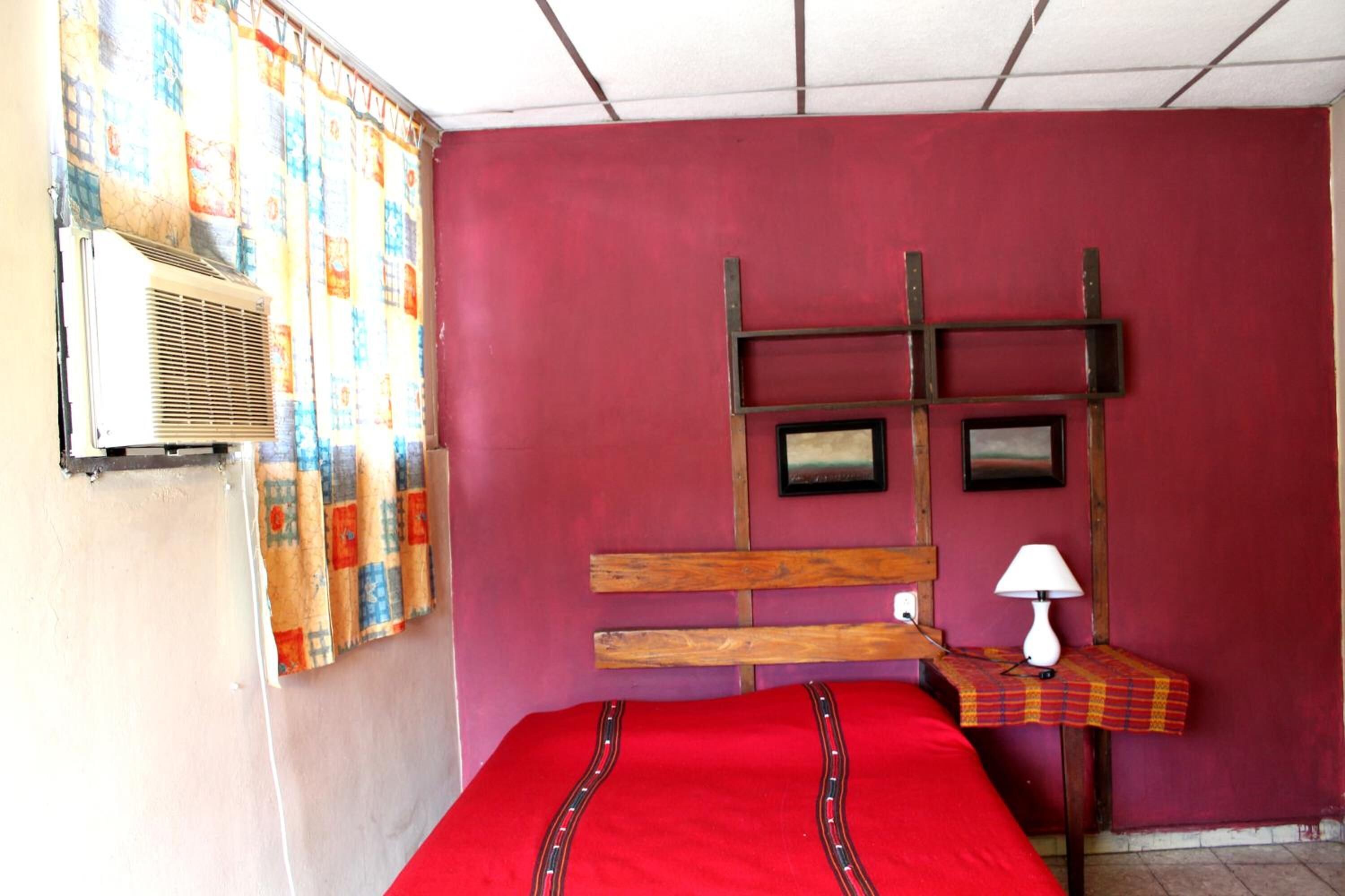 Una stanza con un letto rosso, una testiera in legno, un tavolino con una lampada e un condizionatore d'aria a parete.