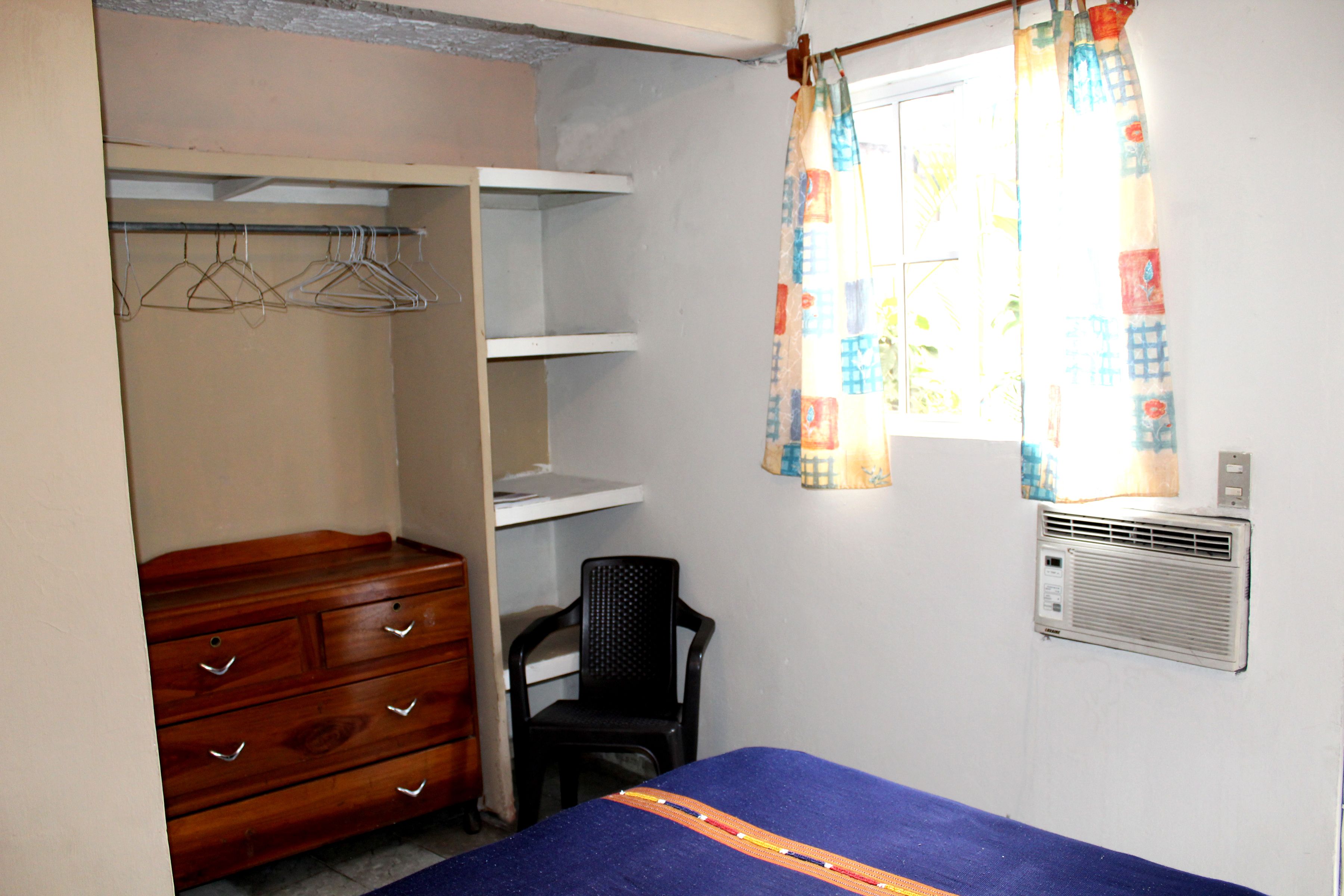 A bedroom with a bed, a wooden dresser, a chair, a wardrobe, and a window with a curtain.