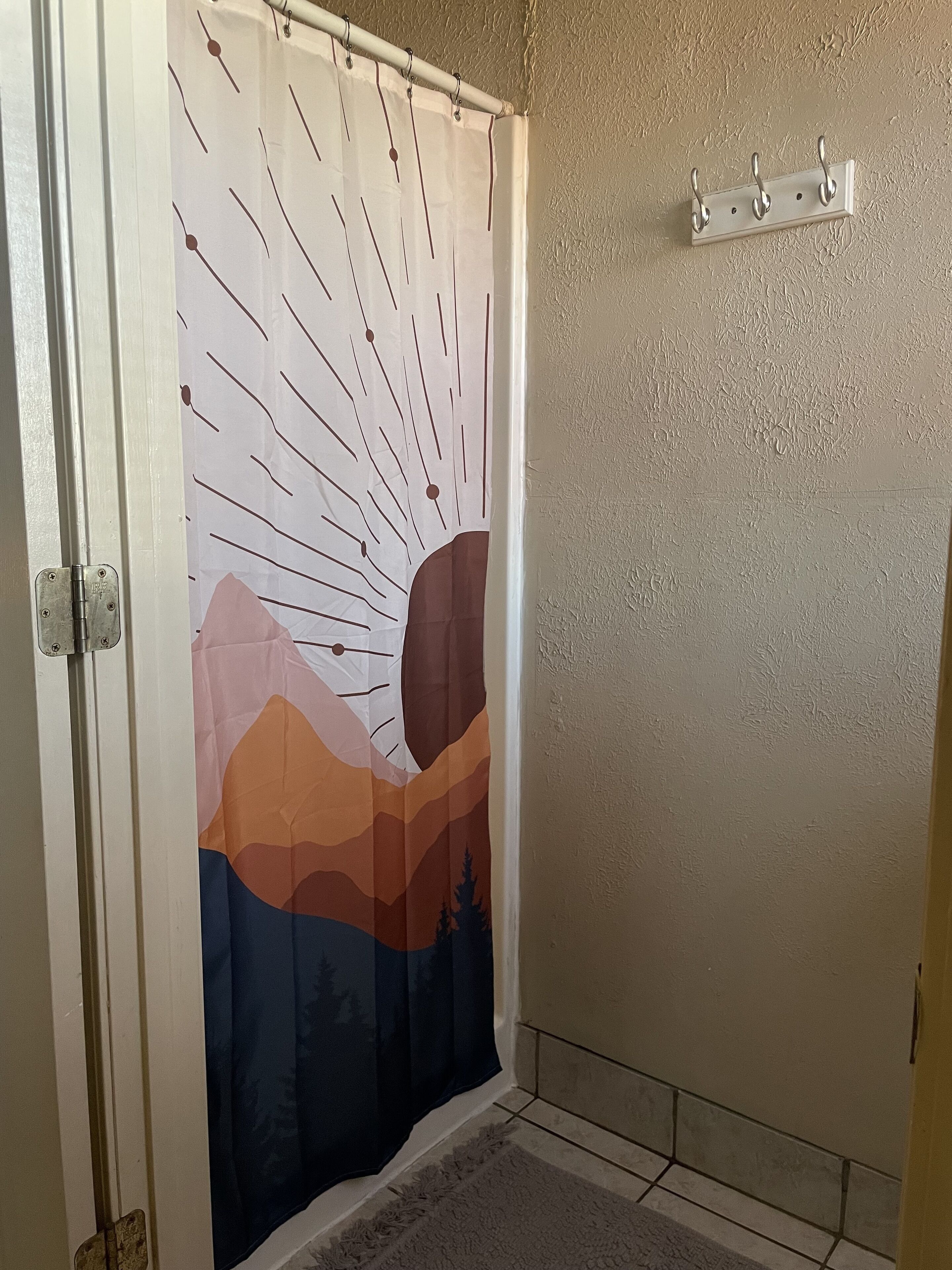 Une salle de bain avec un rideau de douche orné d’un motif représentant un paysage de montagne, un porte-serviettes fixé au mur et un sol carrelé.