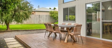 An outdoor patio with a wooden deck, a dining table with wicker chairs, and a view of a modern house with large windows.