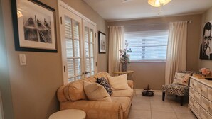 A living room with a beige sofa, a small round table, a patterned armchair, a dresser, and a window with blinds. - Bayshore Inn & Docks Two Bedroom Unit 8 Overlooking the Pool (Naples)