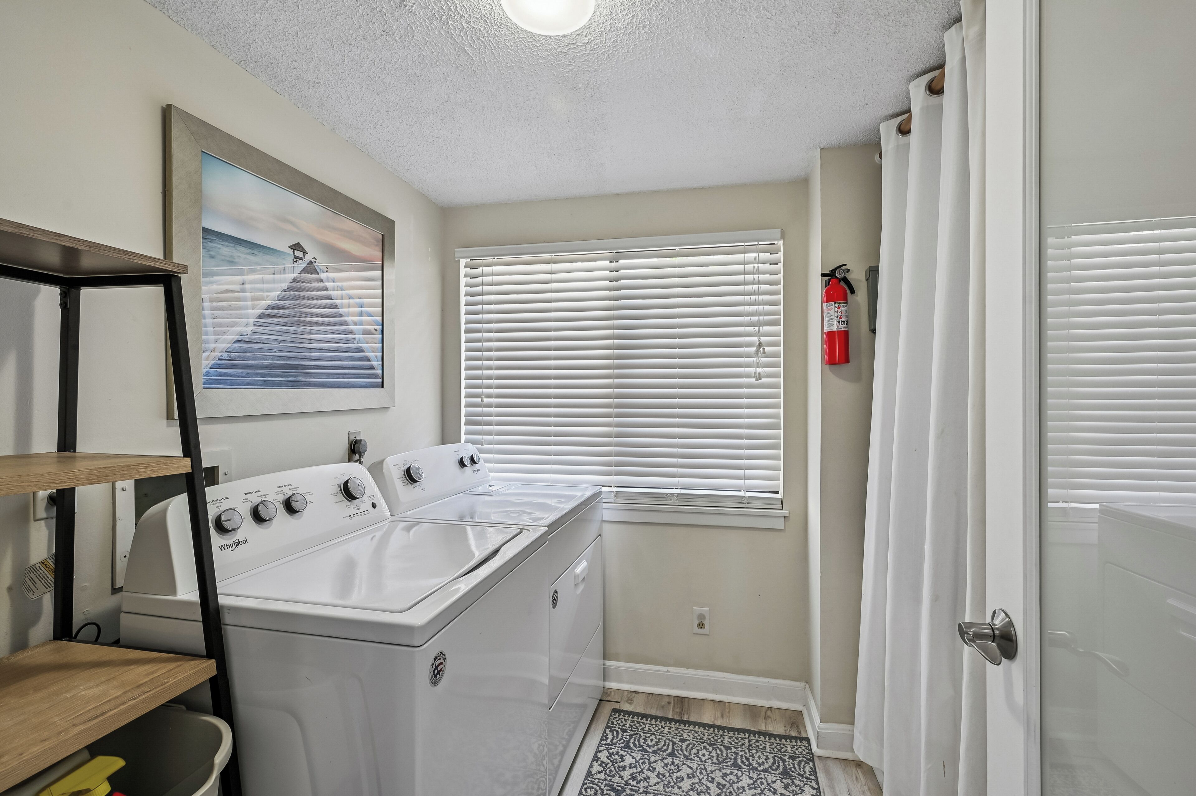 A laundry room with a Whirlpool washer and dryer, a window with blinds, a fire extinguisher, and a door.