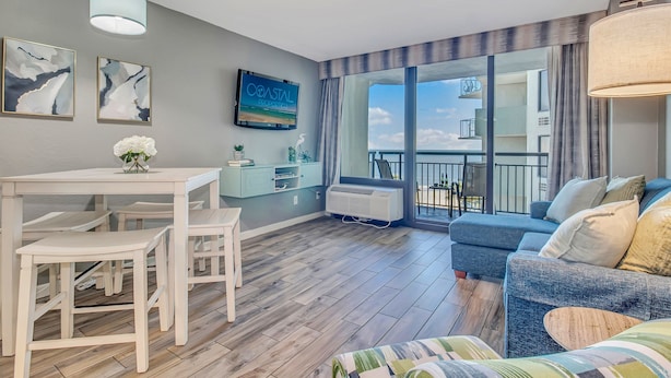 A modern living room with a large sofa, a dining table with chairs, a TV mounted on the wall, and a balcony with a view of the sea.
