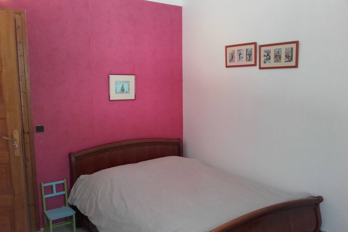 Ein Schlafzimmer mit einem hölzernen Bett, einem grünen Sessel, einer rosa Wand und zwei gerahmten Bildern an der Wand.