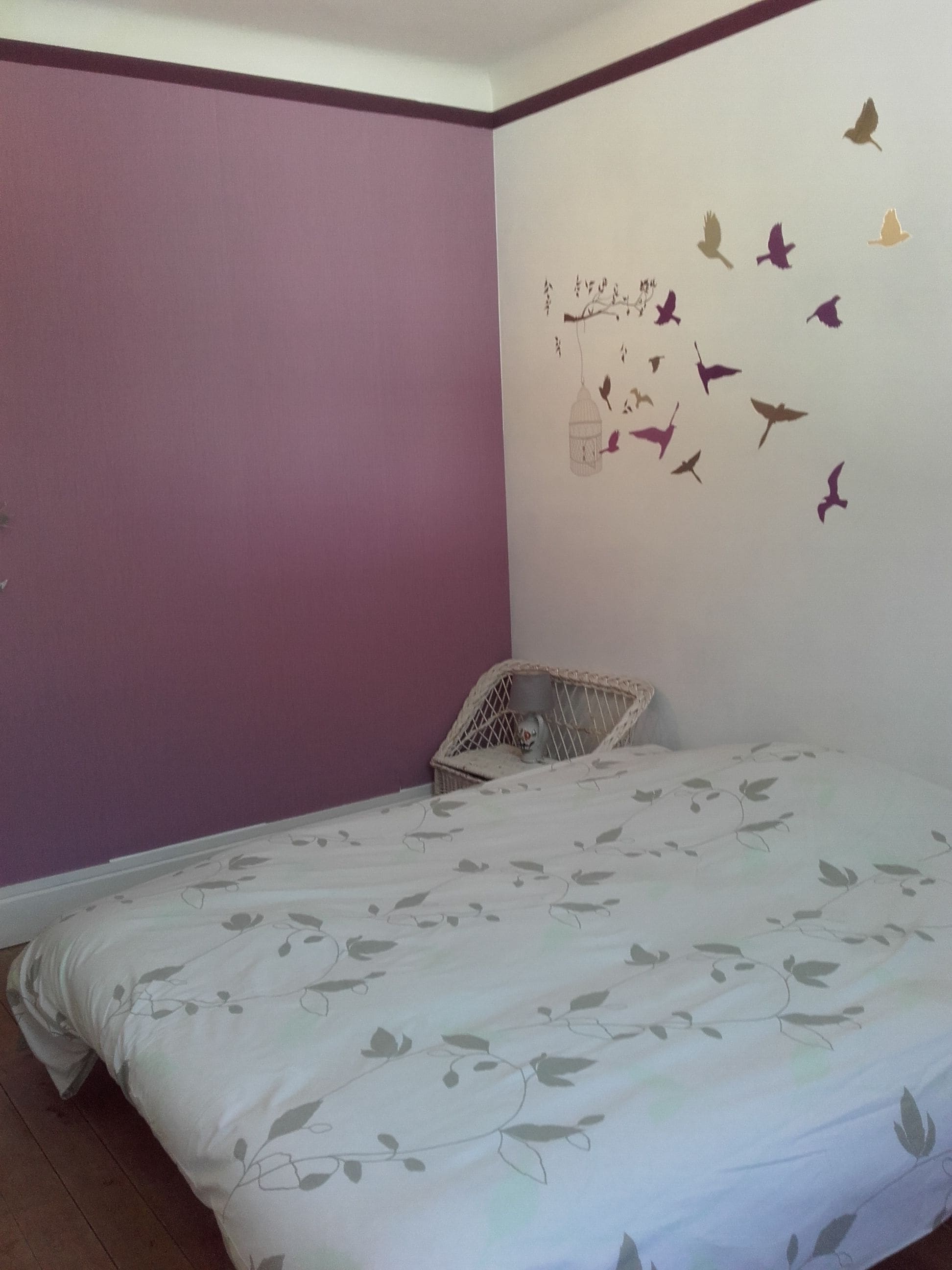 A bedroom with a bed, a wicker chair, and a wall decal of birds.