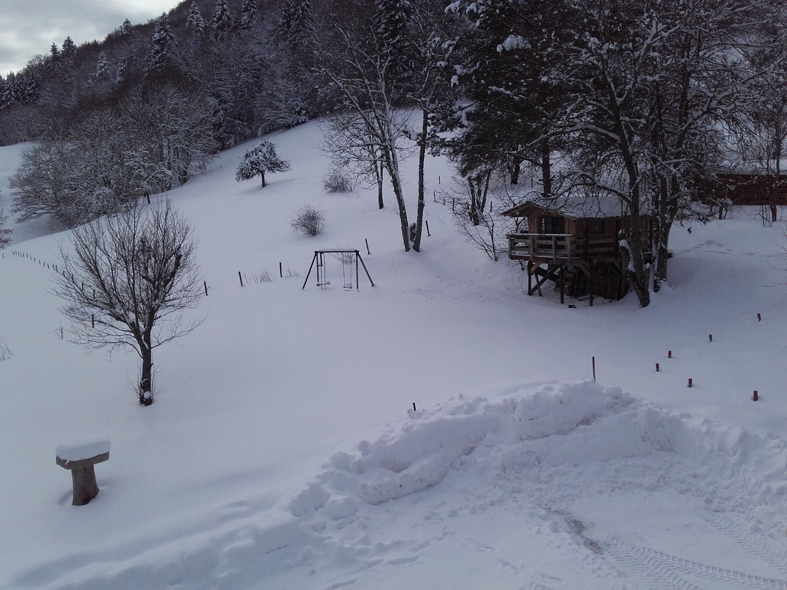 A snow-covered landscape with a wooden playhouse on stilts, a swing set, and a fenced area.