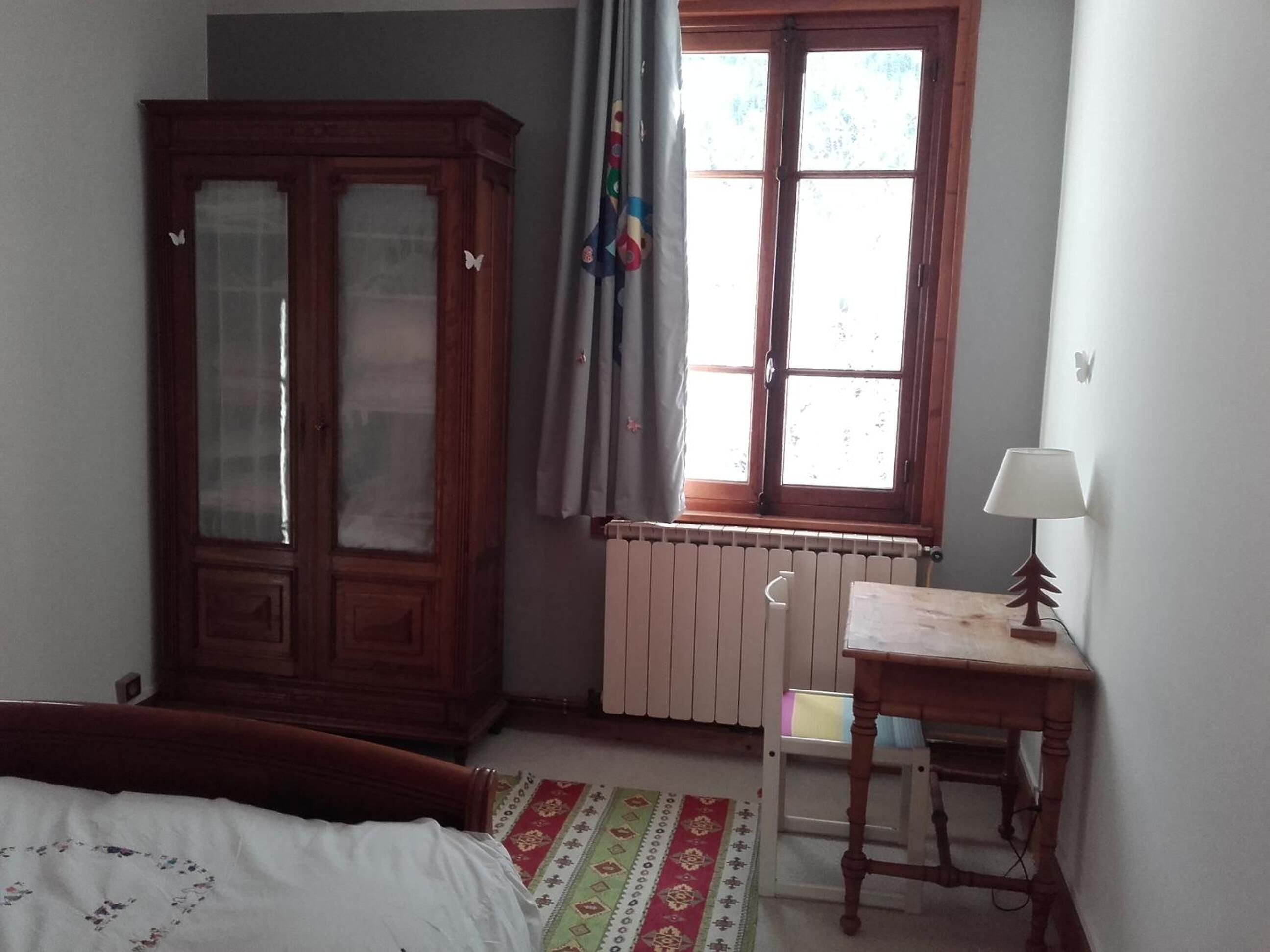 Et soveværelse med en seng, et træskab, en radiator, et vindue med gardiner og et lille bord med en lampe.