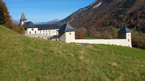Un bâtiment historique doté de nombreuses tours et d’un clocher d’église imposant, entouré d’un paysage verdoyant luxuriant et avec des montagnes en arrière-plan.