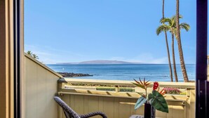 Ein Balkon mit Korbstühlen, einem kleinen Tisch und einem Blumentopf. Blick auf das Meer und Palmen.