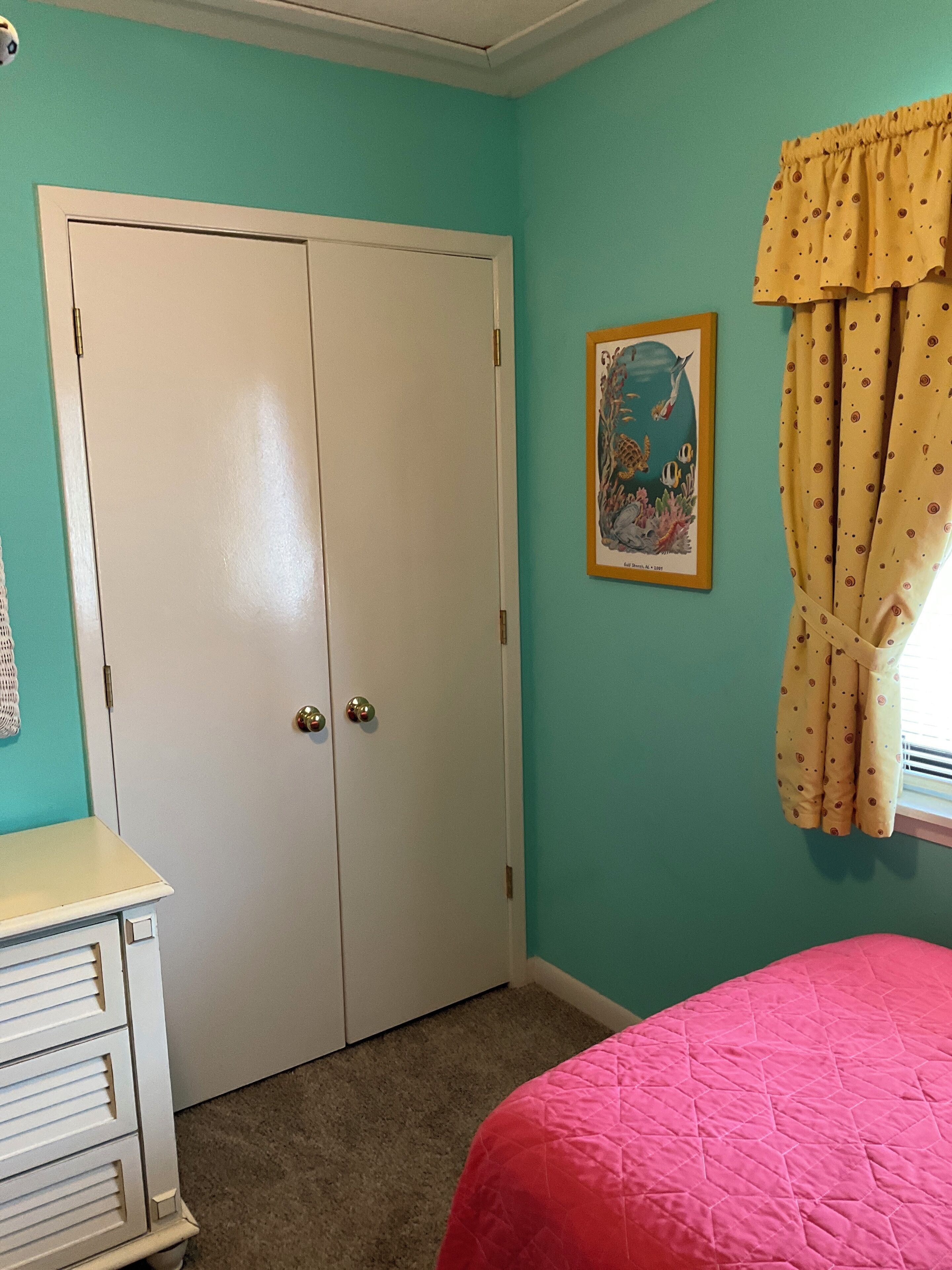 Una habitación con paredes color turquesa, una puerta blanca con dos manijas y un cuadro enmarcado en la pared, además de una ventana con cortinas amarillas.