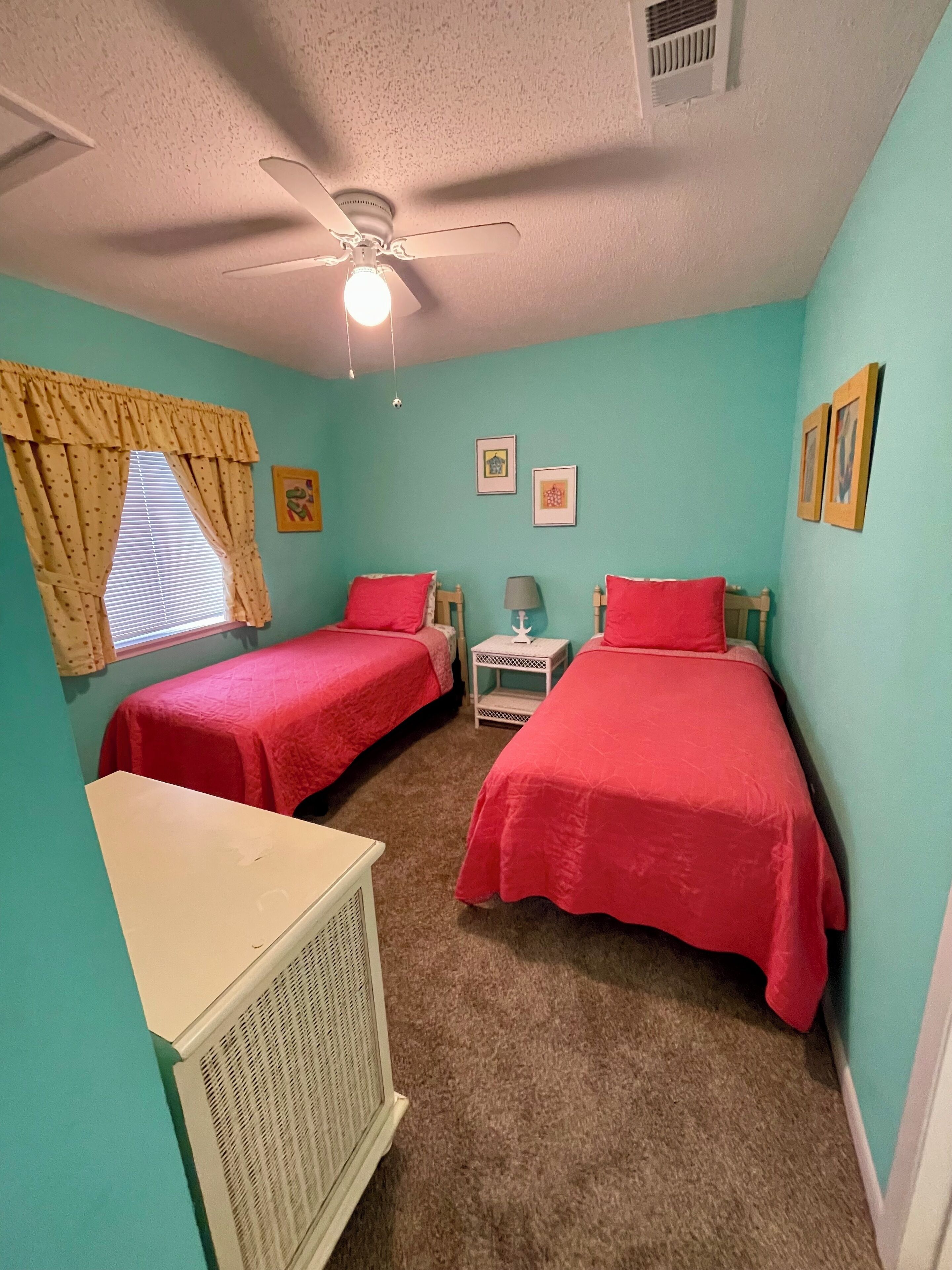 A bedroom with two single beds, a ceiling fan, a radiator, and a window with curtains.