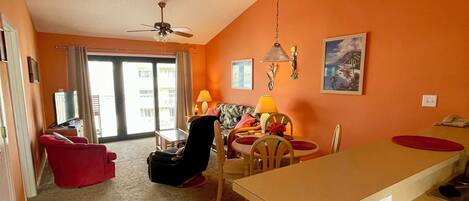 A living room with a red sofa, a dining table with chairs, a ceiling fan, and a painting on the wall.