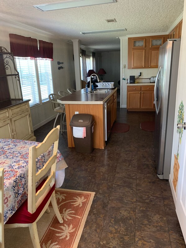 A compact kitchen with wooden cabinets, a central island, a dining area with a patterned tablecloth, and a trash bin.