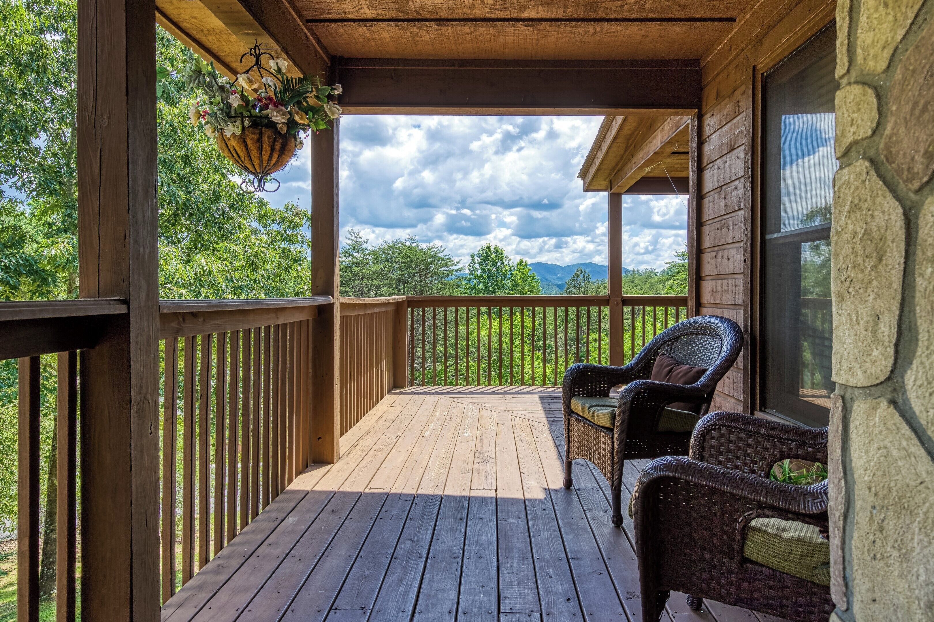 A wooden deck with wicker chairs, a hanging flower basket, and a stone wall.