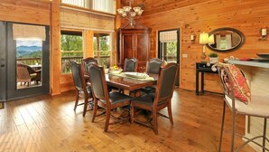 A wooden interior with a dining table and chairs, a chandelier, and a view through a window.