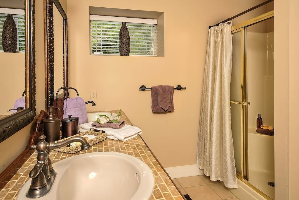 A bathroom with a large mirror, a sink, a window with blinds, a towel rack, and a shower area.