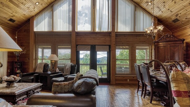 A spacious living room with a high ceiling, wooden beams, a fireplace, a dining area with a table and chairs, and large windows offering a view of the outdoors.