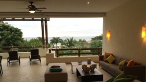 Un vaste espace de vie avec un balcon donnant sur une plage et des palmiers. On y trouve deux canapés, une table basse et des fauteuils.